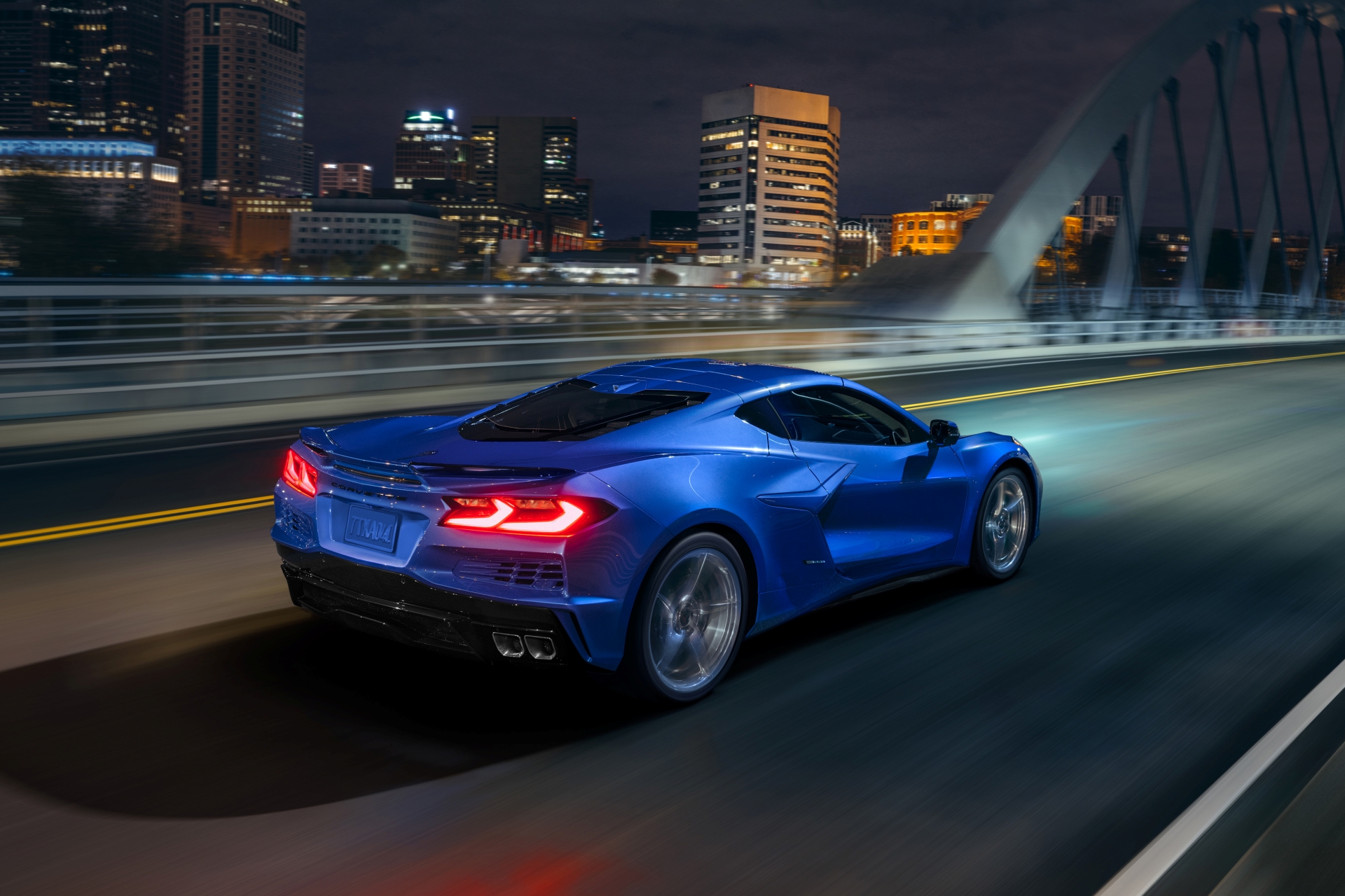 Rear 3/4 view of 2024 Chevrolet Corvette E-Ray 3LZ coupe in Riptide Blue, driving over a bridge into a city. Pre-production model shown. Actual production model may vary. Model year 2024 Corvette E-Ray available 2023.