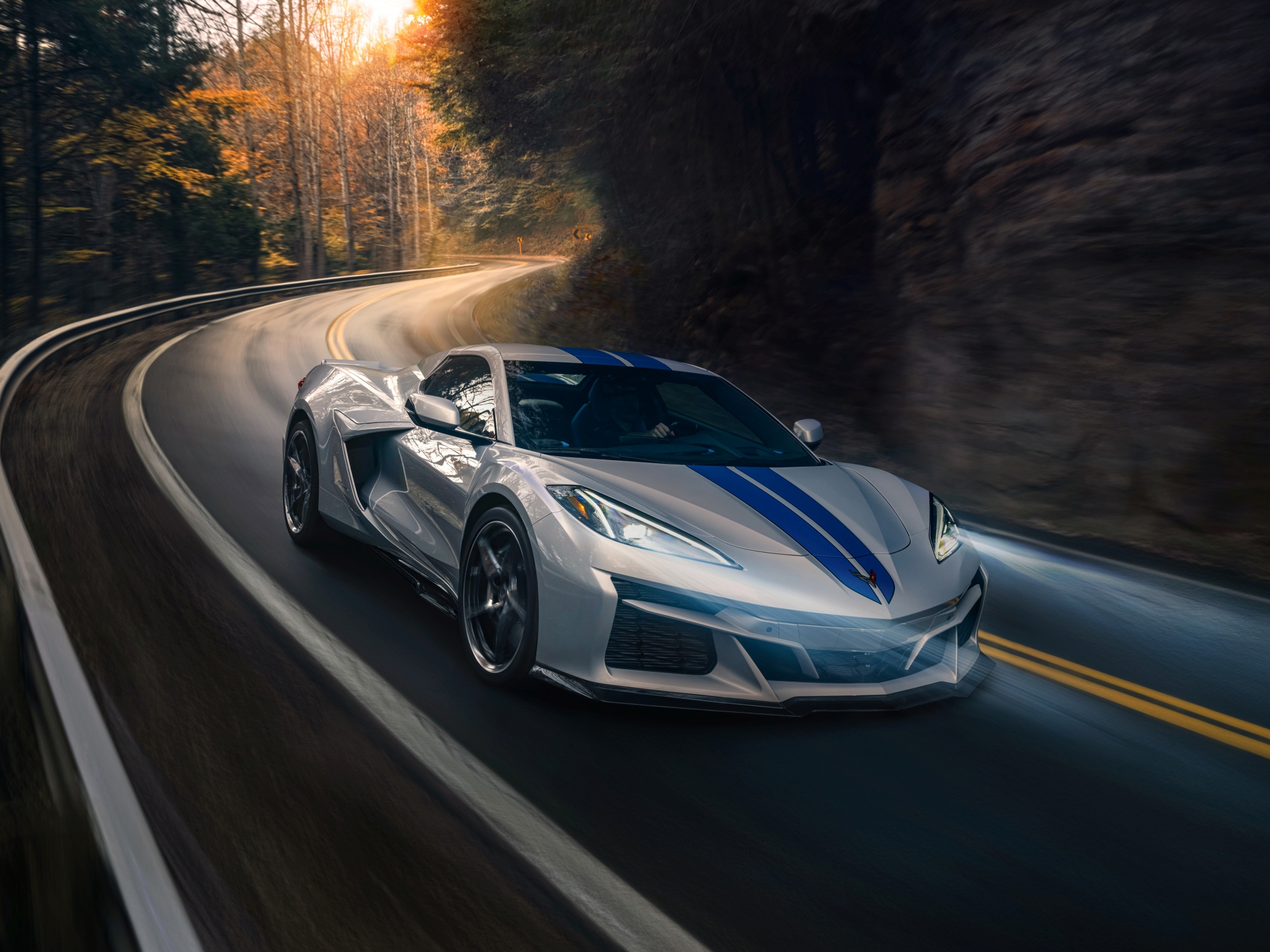 Passenger side front 3/4 view of 2024 Chevrolet Corvette E-Ray 3LZ convertible in Silver Flare with Electric Blue stripe package driving on a curved mountain road. Pre-production model shown. Actual production model may vary. Model year 2024 Corvette E-Ray available 2023.