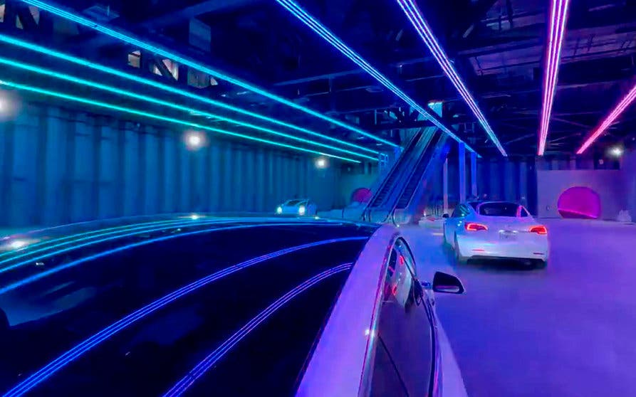  Primera imagen de la estación de pasajeros de Las Vegas Loop bajo luces led multicolor, en la que se aprecian las escaleras mecánicas de acceso, tres Tesla blancos y las bocas de los túneles de entrada y salida. 